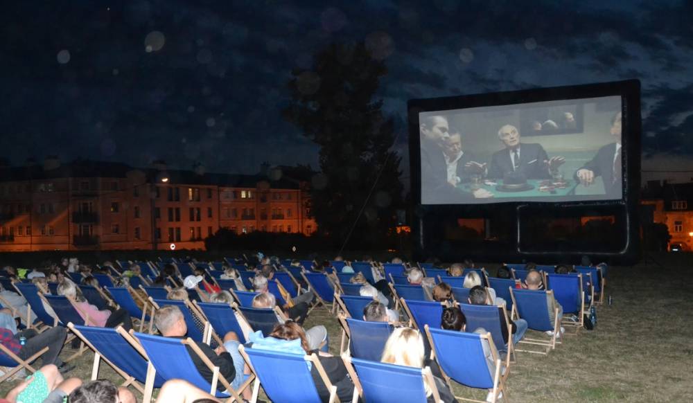 Ludzie siedzący na leżakach oglądają film na dużym telebimie. Jest wieczór, w oddali widać oświetlone budynki.