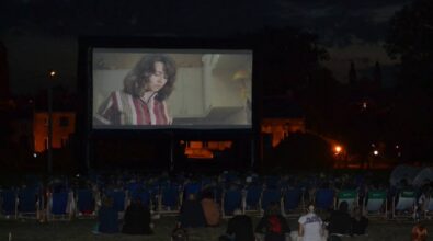 Ludzie siedzący na leżakach oglądają film na dużym telebimie. Jest wieczór, w oddali widać oświetlone budynki. Parę osób siedzi na trawie.