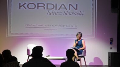 Kobieta siedząca na krześle na scenie, trzyma mikrofon i czyta fragment 