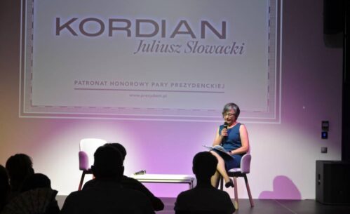 Kobieta siedząca na krześle na scenie, trzyma mikrofon i czyta fragment "Kordiana". W tle widać duży ekran z tekstem "KORDIAN Juliusz Słowacki". Poniżej znajduje się dodatkowy tekst: "PATRONAT HONOROWY PARY PREZYDENCKIEJ". Przed sceną siedzi publiczność.