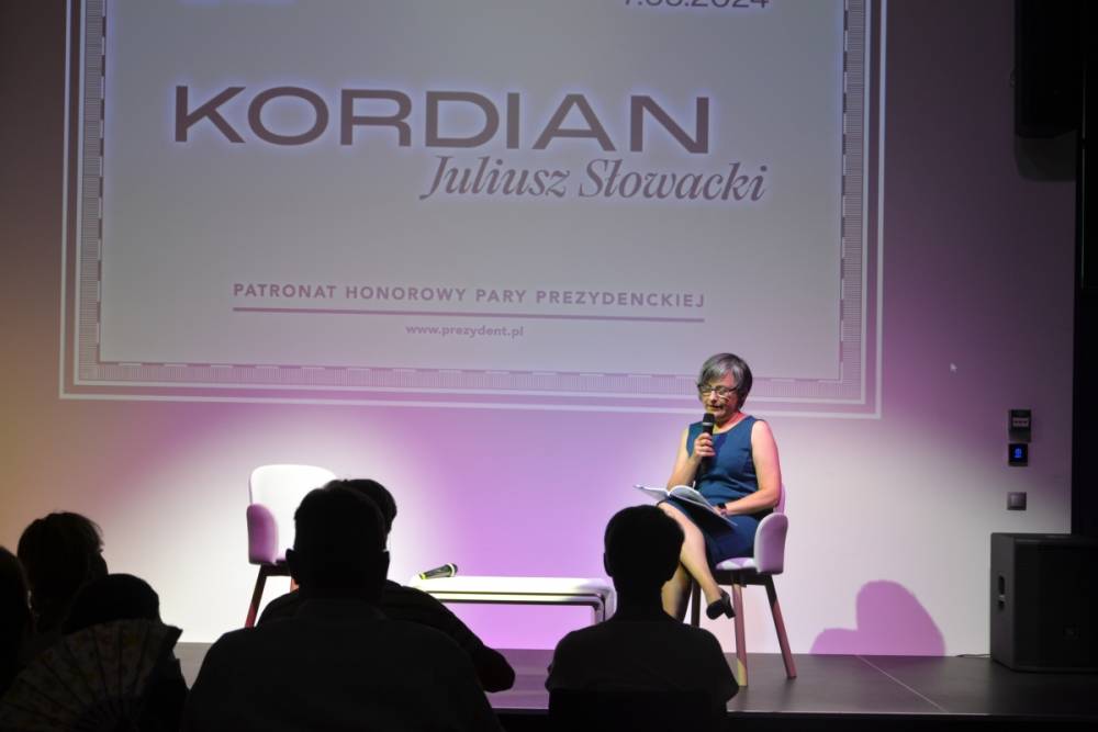 Kobieta siedząca na krześle na scenie, trzyma mikrofon i czyta fragment "Kordiana". W tle widać duży ekran z tekstem "KORDIAN Juliusz Słowacki". Poniżej znajduje się dodatkowy tekst: "PATRONAT HONOROWY PARY PREZYDENCKIEJ". Przed sceną siedzi publiczność.