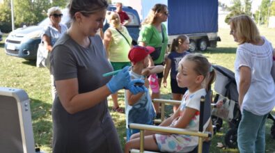 Zdjęcie w plenerze przedstawia uśmiechniętą panią animatorkę, która w rękawiczkach i z pędzelkiem maluje twarz dziecku. Mała dziewczynka siedzi na wysokim krześle. W tle widać dorosłych i dzieci czekających w kolejce.