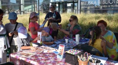 Na zdjęciu widzimy grupę dzieci stojącą przy stole pokrytym kwiecistym obrusem,na którym znajdują się papiery, długopisy i inne przybory artystyczne. Przy stoliku siedzą dwie panie i malują twarze dzieciom. W tle widoczny jest budynek Mediateki.