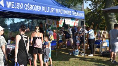 Grupa ludzi zgromadzona pod niebieskim namiotem z napisem ‘MIEJSKI OŚRODEK KULTURY W PIOTRKOWIE TRYBUNALSKIM’. Dzieci i dorośli biorą udział w różnych aktywnościach, takich jak rysowanie czy oglądanie wystawionych przedmiotów. Atmosfera jest żywa i wspólnotowa