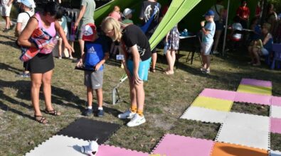 Dziecko stoi na trawie i steruje małym robotem poruszającym się po kolorowych matach ułożonych na ziemi. Obok dwie dorosłe osoby pochylają się i pokazują, jak obsługiwać urządzenie, wskazując kolejne ruchy. Wokół widać inne dzieci i opiekunów korzystających z atrakcji, a w tle znajduje się zielony namiot, pod którym odbywają się zajęcia. Całość ma charakter plenerowych warsztatów i zabawy edukacyjnej.