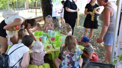 Dzieci siedzą przy niskim stoliku, które wykonują prace plastyczne. Na stolikach znajdują się zielone pojemniki z materiałami. Dzieci ozdabiają papierową koronę. W tle można zauważyć dorosłych, którzy pomagają dzieciom lub obserwują ich zabawę. Niektóre dzieci mają pomalowane twarze. Wokół stolika panuje swobodna atmosfera, a w tle widoczne są drzewa. Z