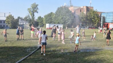 Dzieci i dorośli bawią się na otwartej, trawiastej przestrzeni, gdzie straż pożarna zorganizowała wodną kurtynę. Dzieci z radością biegają i skaczą w strumieniach wody, korzystając z ochłody w gorący dzień. W tle widać budynki i drzewka, a także strażacki wóz. Uczestnicy imprezy wyglądają na bardzo zadowolonych, a atmosfera jest pełna zabawy i beztroski.