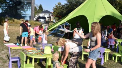 Na trawie ustawione są małe stoliki i krzesełka dla dzieci, przy których najmłodsi bawią się klockami i różnymi akcesoriami. W centrum jedna z osób tworzy duże bańki mydlane, które unoszą się nad stolikiem i przyciągają uwagę dzieci. Obok dorosła osoba pomaga dziecku sięgnąć po zabawkę z pojemnika. W tle znajduje się zielony namiot, pod którym siedzą uczestnicy wydarzenia, a dalej widać grupy dzieci i opiekunów korzystających z przygotowanych atrakcji na świeżym powietrzu.