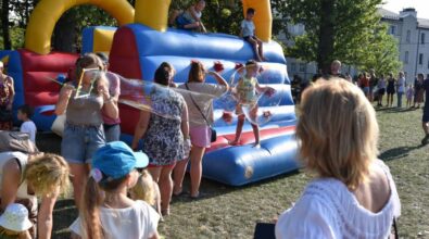Dzieci i dorośli bawią się na trawie przy dużej dmuchanej zjeżdżalni w kolorach niebieskim, czerwonym i żółtym. Kilkoro dzieci znajduje się na górze i zjeżdża, inne stoją na dole lub czekają na swoją kolej. Wśród uczestników jedna z osób tworzy duże bańki mydlane, które unoszą się w powietrzu i przyciągają uwagę dzieci. Wokół zgromadzone są rodziny obserwujące zabawę, a całość odbywa się w słonecznej, plenerowej atmosferze.dmuchana konstrukcja, na której bawią się dzieci. Atmosfera jest pełna życia, a ludzie cieszą się słoneczną pogodą na zielonej, otwartej przestrzeni