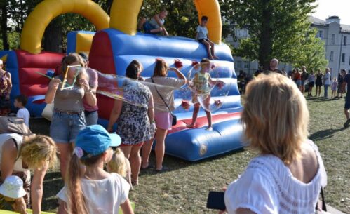 Dzieci i dorośli bawią się na trawie przy dużej dmuchanej zjeżdżalni w kolorach niebieskim, czerwonym i żółtym. Kilkoro dzieci znajduje się na górze i zjeżdża, inne stoją na dole lub czekają na swoją kolej. Wśród uczestników jedna z osób tworzy duże bańki mydlane, które unoszą się w powietrzu i przyciągają uwagę dzieci. Wokół zgromadzone są rodziny obserwujące zabawę, a całość odbywa się w słonecznej, plenerowej atmosferze.dmuchana konstrukcja, na której bawią się dzieci. Atmosfera jest pełna życia, a ludzie cieszą się słoneczną pogodą na zielonej, otwartej przestrzeni