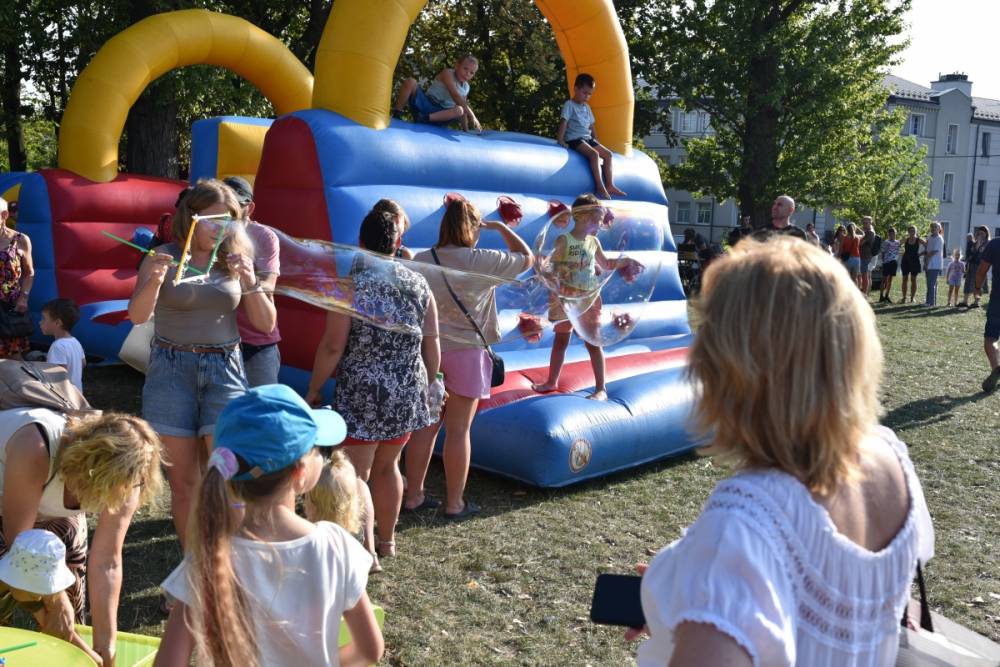 Dzieci i dorośli bawią się na trawie przy dużej dmuchanej zjeżdżalni w kolorach niebieskim, czerwonym i żółtym. Kilkoro dzieci znajduje się na górze i zjeżdża, inne stoją na dole lub czekają na swoją kolej. Wśród uczestników jedna z osób tworzy duże bańki mydlane, które unoszą się w powietrzu i przyciągają uwagę dzieci. Wokół zgromadzone są rodziny obserwujące zabawę, a całość odbywa się w słonecznej, plenerowej atmosferze.dmuchana konstrukcja, na której bawią się dzieci. Atmosfera jest pełna życia, a ludzie cieszą się słoneczną pogodą na zielonej, otwartej przestrzeni