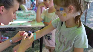 Na pierwszym planie pani animatorka robi tatuaż na ręce dziecka. W tle widać stolik z szablonami tatuaży oraz grupkę dzieci.
