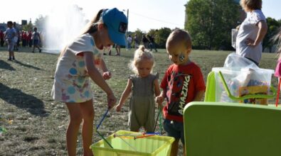Troje małych dzieci stoi na trawie i bawi się przy pojemniku z wodą, używając kolorowych rurek lub patyczków do zabawy. Dziewczynka w czapce pochyla się nad pojemnikiem, a dwoje młodszych dzieci stoi obok i obserwuje, trzymając elementy zabawki. Obok znajduje się zielony stolik z pudełkiem pełnym akcesoriów. W tle widać inne osoby uczestniczące w wydarzeniu oraz fontannę wodną, a całość odbywa się na świeżym powietrzu w słoneczny dzień.