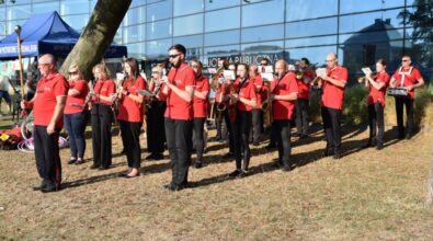 Grupa muzyków ubranych w czerwone koszulki stoi na trawie przed przeszklonym budynkiem Mediateki i gra na instrumentach dętych. Wśród nich są osoby grające na saksofonach, trąbkach i klarnecie, trzymające nuty na pulpitach lub w rękach. W tle widoczny jest namiot oraz elementy wydarzenia plenerowego, a całość odbywa się w słonecznej pogodzie, wśród zieleni i drzew.