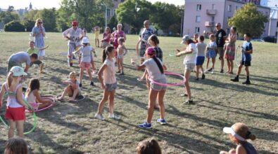 Grupa dzieci bawi się na trawie na otwartej przestrzeni, kręcąc kolorowymi hula-hopami. Niektóre dzieci stoją i próbują utrzymać obręcze w ruchu, inne siedzą lub rozmawiają, trzymając je w rękach. Wokół stoją dorośli opiekunowie, którzy obserwują zabawę. W tle widoczne są budynki oraz drzewa, a całość odbywa się w słonecznej, plenerowej atmosferze.