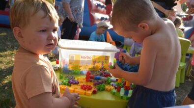 Dwoje małych dzieci stoi przy niskim stoliku i bawi się kolorowymi klockami konstrukcyjnymi. Jedno z nich trzyma element i uważnie go dopasowuje, drugie spogląda w bok, trzymając w dłoni część zabawki. Na stole znajduje się pudełko z dodatkowymi klockami oraz rozłożone elementy w różnych kolorach i kształtach. W tle widoczne są inne dzieci oraz dmuchana atrakcja, co wskazuje na plenerową zabawę lub wydarzenie dla najmłodszych.