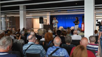 Widok na salę wypełnioną publicznością, głównie starszymi osobami, siedzącymi na krzesłach i słuchającymi wystąpienia na scenie. Na scenie znajduje się starszy mężczyzna w białym stroju, trzymający dokumenty, oraz młodsza kobieta w ciemnozielonej bluzce i białych spodniach, która prowadzi wydarzenie z mikrofonem w ręku. Za nimi widoczna jest niebieska ściana. Z lewej strony znajduje się głośnik, a w tle dostrzec można półki z książkami. Po prawej stronie stoi kamerzysta, który rejestruje całe wydarzenie.