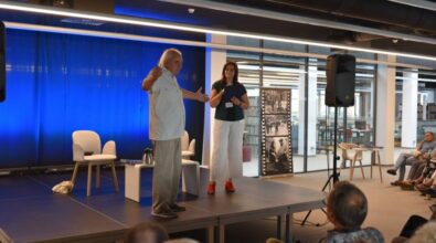 Na zdjęciu widać dwie osoby na scenie podczas wydarzenia kulturalnego. Starszy mężczyzna z siwymi włosami, ubrany w jasny strój, gestykuluje rękoma, jakby opowiadał coś publiczności. Obok niego stoi kobieta w ciemnozielonej bluzce i białych spodniach, trzymająca niebieską teczkę i uważnie słuchająca mówcy. Scena jest oświetlona i ma tło z niebieską kotarą. Na scenie znajdują się białe krzesła i stolik z dzbankiem oraz książkami. W tle widoczne są duże przeszklenia, przez które można dostrzec regały z książkami. Przed sceną siedzą uczestnicy spotkania, a po prawej stronie na krzesłach z boku widać kilka osób, które także obserwują wystąpienie.