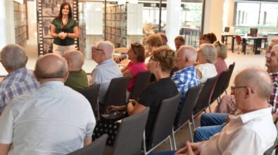 Widok na salę wypełnioną publicznością , siedzącą na krzesłach. Po prawej stronie stoi młoda kobieta, ubrana w ciemnozieloną bluzkę i białe spodnie. W tle widać półki z książkami
