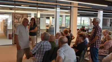 Grupa osób uczestnicząca w spotkaniu w bibliotece. Jeden z uczestników stoi i zabiera głos, a reszta publiczności siedzi na krzesłach, aktywnie słuchając. Prowadząca z mikrofonem stoi przed grupą, na tle tablicy z archiwalnymi zdjęciami. W tle widoczne są regały z książkami oraz nowoczesna przestrzeń biblioteczna. Atmosfera wydarzenia sprzyja dialogowi i integracji uczestników.