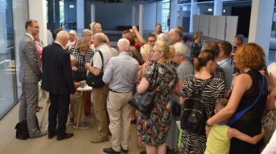 Grupa osób zebranych w jasnym, nowoczesnym wnętrzu biblioteki. W centrum zdjęcia stoi kilka osób, które prowadzą rozmowę i uważnie przyglądają się osobie siedzącej za stolikiem. Wokół zgromadziło się kilkanaście osób w różnym wieku, w większości seniorzy, ubrane elegancko i w letnie stroje. Na pierwszym planie kobieta w kolorowej sukience i okularach trzyma dużą czarną torebkę, obok niej kobieta z dzieckiem ubranym na jasnozielono. W tle widoczne są inne osoby, wnętrze z dużymi oknami oraz przestrzeń wystawowa z białymi panelami. W grupie panuje przyjazna, żywiołowa atmosfera.