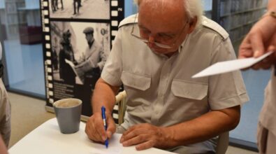 Jerzy Rogalski siedzi przy stole, podpisując kartkę długopisem. Ubrany jest w jasną koszulę z krótkim rękawem. Przed nim stoi szary kubek z napojem, a obok leży niebieski długopis. W tle widoczna jest wystawa ze zdjęciami historycznymi, przedstawiającymi sceny z życia codziennego. Po prawej stronie ktoś trzyma kartkę papieru, prawdopodobnie czekając na podpis lub dedykację. Cała scena ma miejsce w pomieszczeniu.