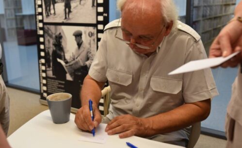 Jerzy Rogalski siedzi przy stole, podpisując kartkę długopisem. Ubrany jest w jasną koszulę z krótkim rękawem. Przed nim stoi szary kubek z napojem, a obok leży niebieski długopis. W tle widoczna jest wystawa ze zdjęciami historycznymi, przedstawiającymi sceny z życia codziennego. Po prawej stronie ktoś trzyma kartkę papieru, prawdopodobnie czekając na podpis lub dedykację. Cała scena ma miejsce w pomieszczeniu.