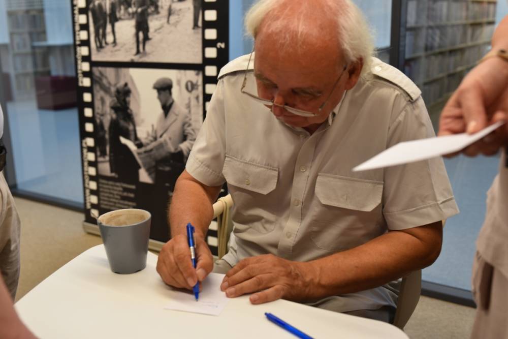 Jerzy Rogalski siedzi przy stole, podpisując kartkę długopisem. Ubrany jest w jasną koszulę z krótkim rękawem. Przed nim stoi szary kubek z napojem, a obok leży niebieski długopis. W tle widoczna jest wystawa ze zdjęciami historycznymi, przedstawiającymi sceny z życia codziennego. Po prawej stronie ktoś trzyma kartkę papieru, prawdopodobnie czekając na podpis lub dedykację. Cała scena ma miejsce w pomieszczeniu.