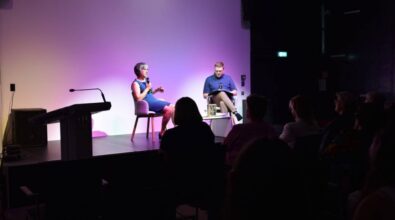 Kobieta i mężczyzna siedzą na scenie i rozmawiają. Przed sceną siedzi publiczność.