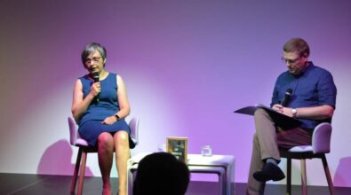 Kobieta i mężczyzna siedzą na scenie i rozmawiają. Przed sceną siedzi publiczność.