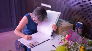 Agnieszka Gehrke siedze przy stoliku i wpisuje się do kroniki biblioteki. Przed nią leży bukiet kwiatów