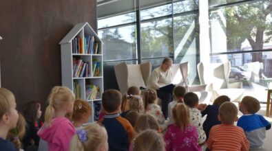 Grupa dzieci siedzi na podłodze przed starszą osobą, która czyta książkę. Pomieszczenie ma duże okna, przez które wpada naturalne światło, oraz kilka nowoczesnych, wygodnie wyglądających krzeseł rozstawionych wokół czytającej osoby. Po lewej stronie widać półkę z różnymi książkami.