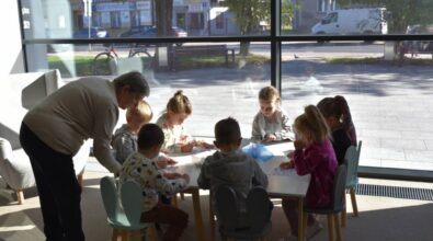 Grupa dzieci siedzi przy stoliku i wykonuje zadanie na kartce. Obok nad stolikiem pochyla się starsza pani.