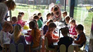 Grupa dzieci siedzi przy stoliku i wykonuje zadanie na kartce. Obok nad stolikiem pochylają się panie.