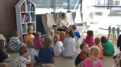 Grupa dzieci siedzi na podłodze przed starszą osobą, która czyta książkę. Pomieszczenie ma duże okna, przez które wpada naturalne światło, oraz kilka nowoczesnych, wygodnie wyglądających krzeseł rozstawionych wokół czytającej osoby. Po lewej stronie widać półkę z różnymi książkami.