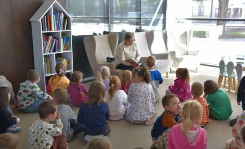 Grupa dzieci siedzi na podłodze przed starszą osobą, która czyta książkę. Pomieszczenie ma duże okna, przez które wpada naturalne światło, oraz kilka nowoczesnych, wygodnie wyglądających krzeseł rozstawionych wokół czytającej osoby. Po lewej stronie widać półkę z różnymi książkami.