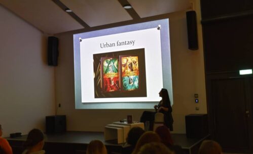 Widok na kolejny slajd prezentacji, osobę prowadzącą oraz publiczność.