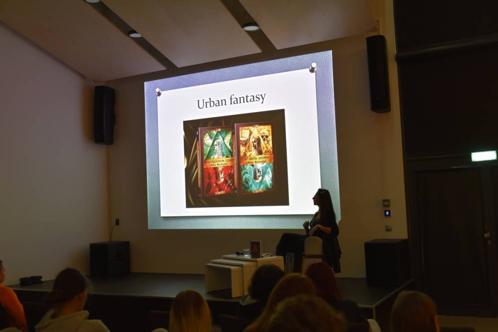 Widok na kolejny slajd prezentacji, osobę prowadzącą oraz publiczność.