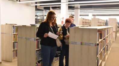 Trzy osoby chodzą między regałami z książkami. Jedna z nich trzyma w ręku telefon, kartkę i ołówek.