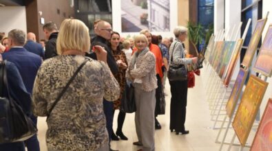 Wernisaż prac malarskich, odbywający się w eleganckim, przestronnym wnętrzu z dużymi oknami. Na sztalugach rozmieszczone są różnorodne obrazy, które przyciągają uwagę zgromadzonych gości. Widzowie, w różnym wieku, przyglądają się eksponatom. W tle widać żywą atmosferę, a światło oświetla zarówno prace, jak i osoby uczestniczące w wydarzeniu.