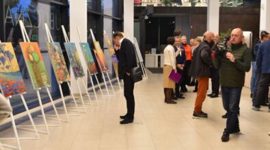 sztalugi z obrazami, osoba w czarnym garniturze z zainteresowaniem ogląda wystawę. Obok widać rozmawiającą grupkę ludzi, a także mężczyznę z kamerą.