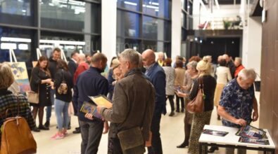 Zgromadzone osoby oglądają wystawę. Niektórzy stoją w grupkach i rozmawiają ze sobą. Parę osób trzyma broszury. Po prawej mężczyzna bierze broszurę ze stolika.