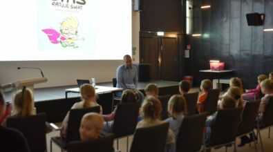 Prowadzący, siedząc na krześle przed grupą dzieci, prowadzi ożywioną rozmowę. W tle widać ekran projekcyjny i kolorowy rysunek.