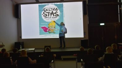 Prowadzący stoi na scenie i pokazuje na ekran projekcyjny. W drugiej ręce trzyma książki. Na ekranie kolorowa okładka jego książki pod tytułem 