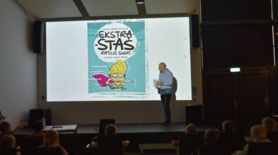 Prowadzący stoi na scenie i patrzy na ekran projekcyjny. W rękach trzyma książki. Na ekranie kolorowa okładka jego książki pod tytułem 