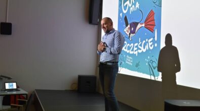 Tomasz Nowaczyk stoi uśmiechnięta, trzymając ręce złożone przed sobą, na tle ekranu projekcyjnego. Na ekranie wyświetla się kolorowa okładka książki z rybą pod tytułem 