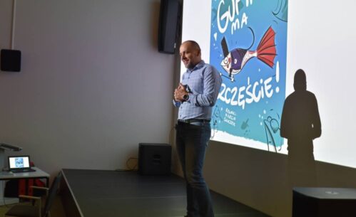 Tomasz Nowaczyk stoi uśmiechnięta, trzymając ręce złożone przed sobą, na tle ekranu projekcyjnego. Na ekranie wyświetla się kolorowa okładka książki z rybą pod tytułem "Gupik ma szczęście".