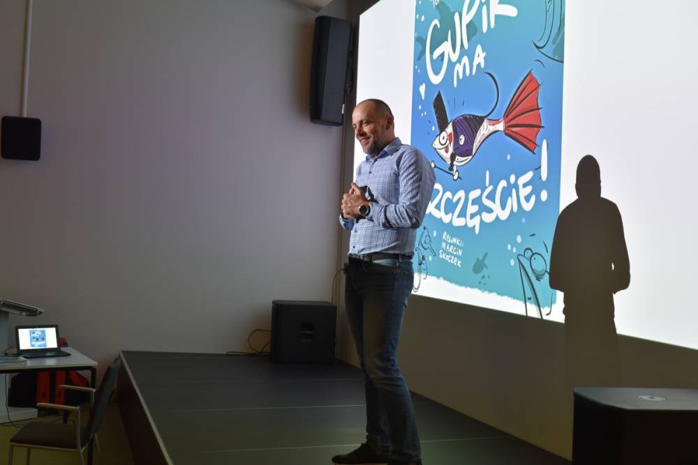 Tomasz Nowaczyk stoi uśmiechnięta, trzymając ręce złożone przed sobą, na tle ekranu projekcyjnego. Na ekranie wyświetla się kolorowa okładka książki z rybą pod tytułem "Gupik ma szczęście".