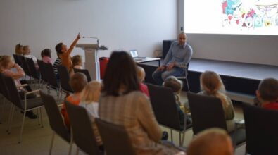 Uśmiechnięty prowadzący siedzi na krześle przed dziećmi i rozmawiam z nimi. Dzieci są zadowolone, niektóre się śmieją.