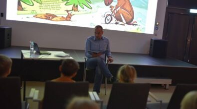 Prowadzący siedzi na krześle przed dziećmi i czyta książkę. W tle na ekranie widać stronę z książki przedstawiającą rysunkowego łosia jadącego na rowerze w lesie.